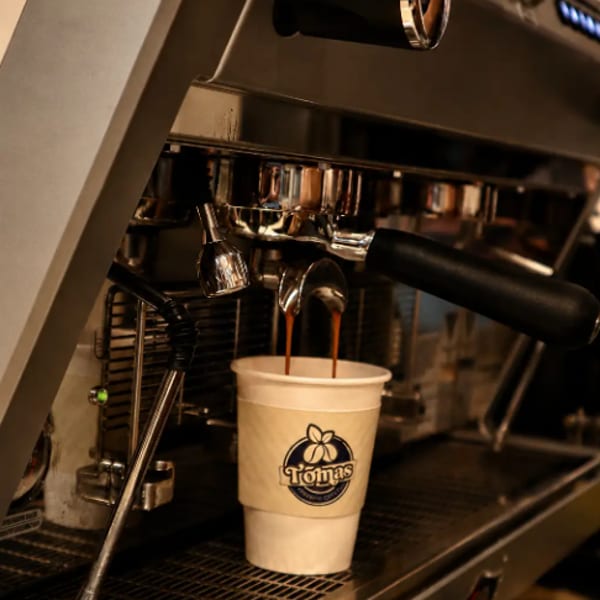 Espresso pouring from a machine into a white paper cup with the Tomas logo.