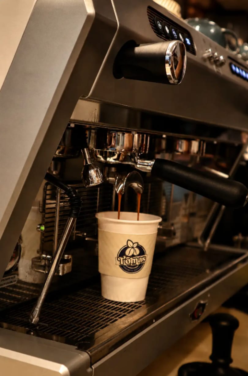Espresso pouring from a machine into a white paper cup with the Tomas logo.